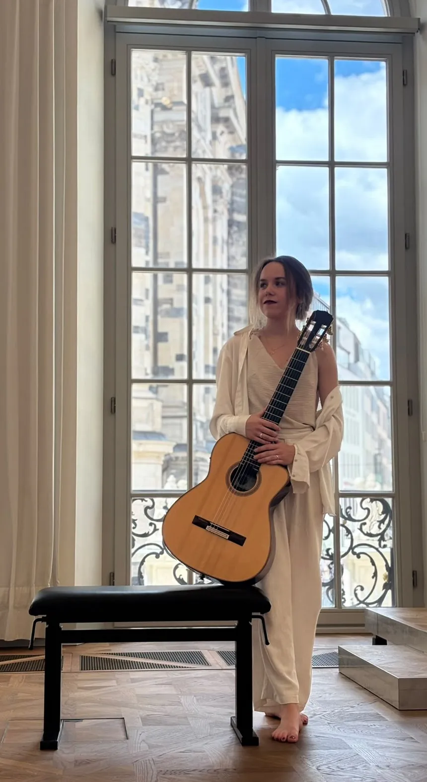 Margaréta Lakner standing with her guitar at a tall window overlooking Dresden